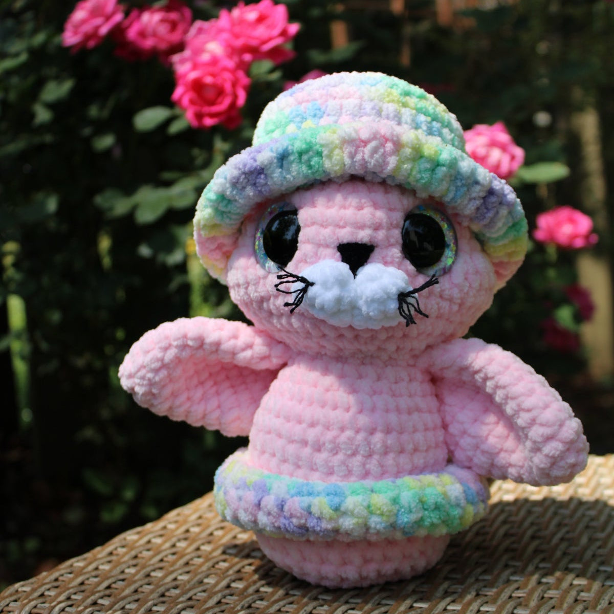Pink crochet seal toy with a colorful hat on a wicker surface with flowers in the background