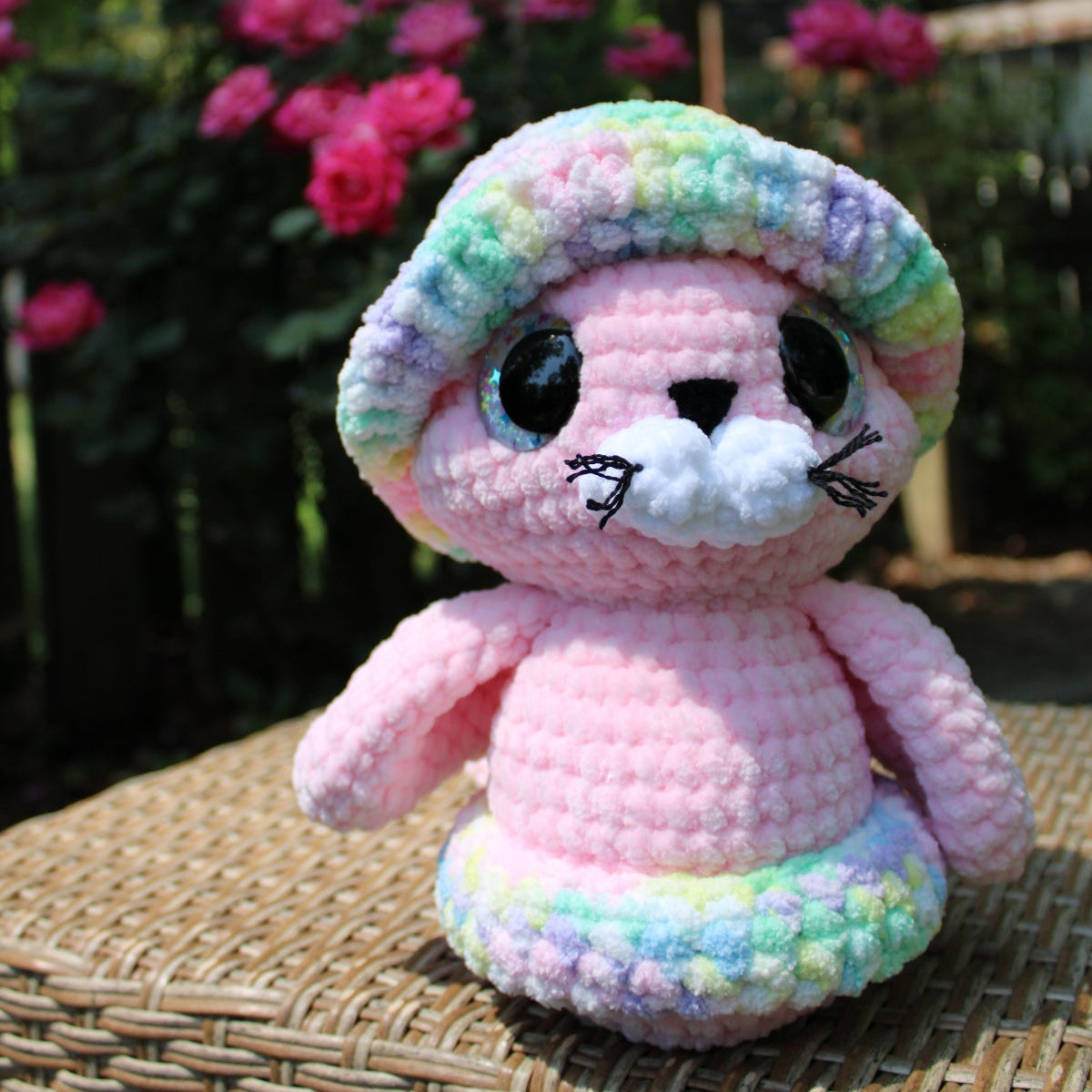 Pink crochet toy with a rainbow hat on a wicker surface with flowers in the background
