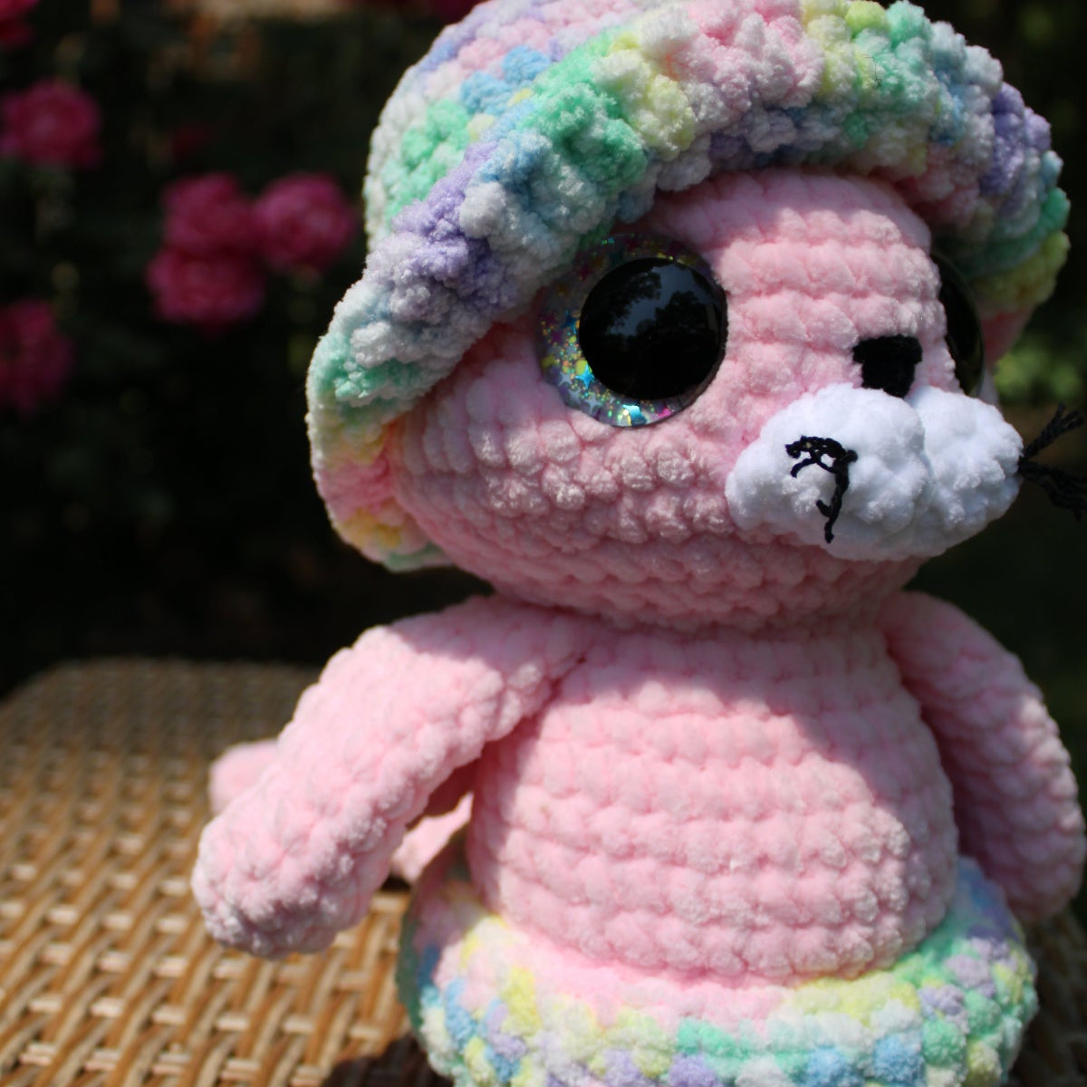 Pink crochet seal with rainbow hat on a wicker surface with blurred flowers in the background
