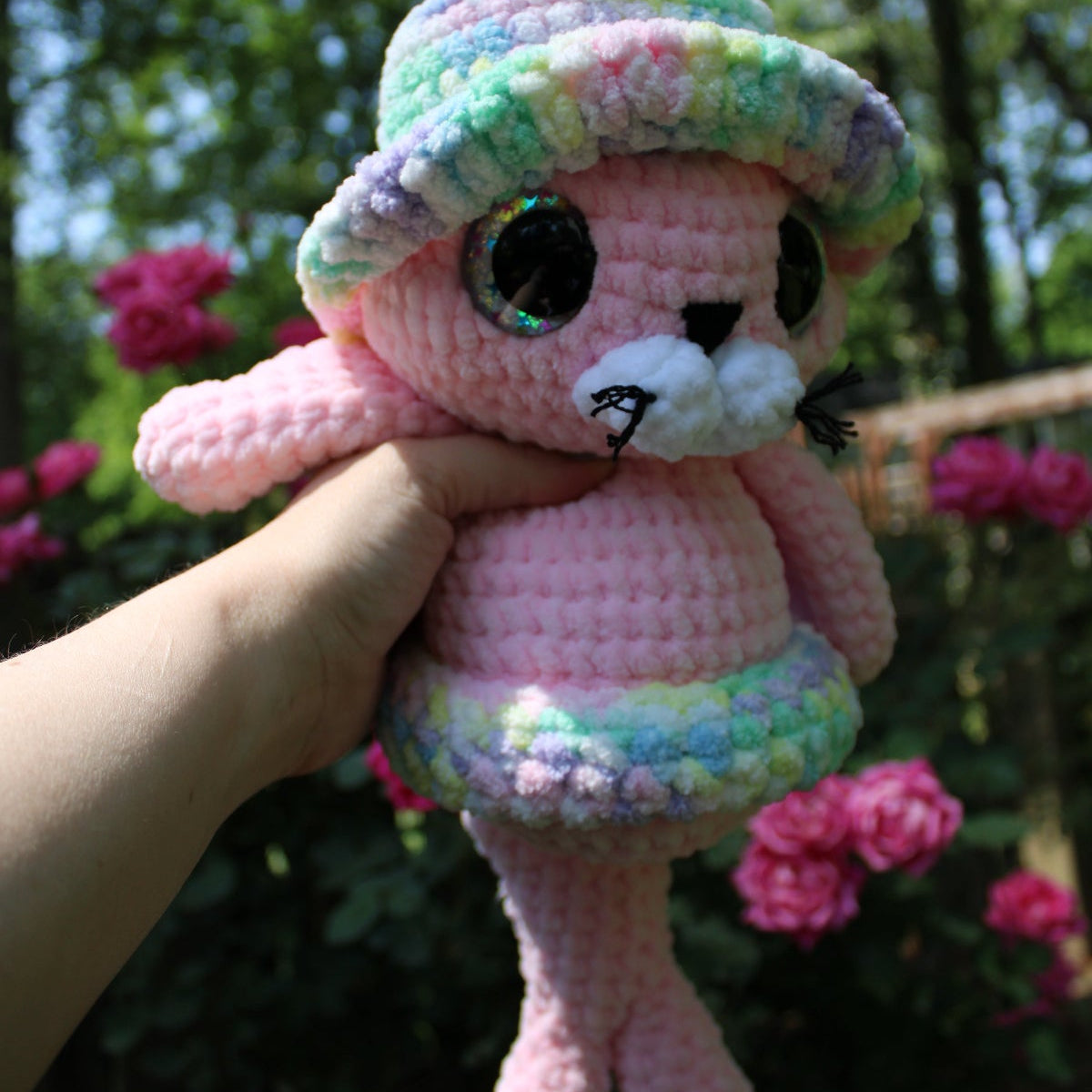 Pink crochet plush toy with a colorful hat held in front of pink flowers and greenery.