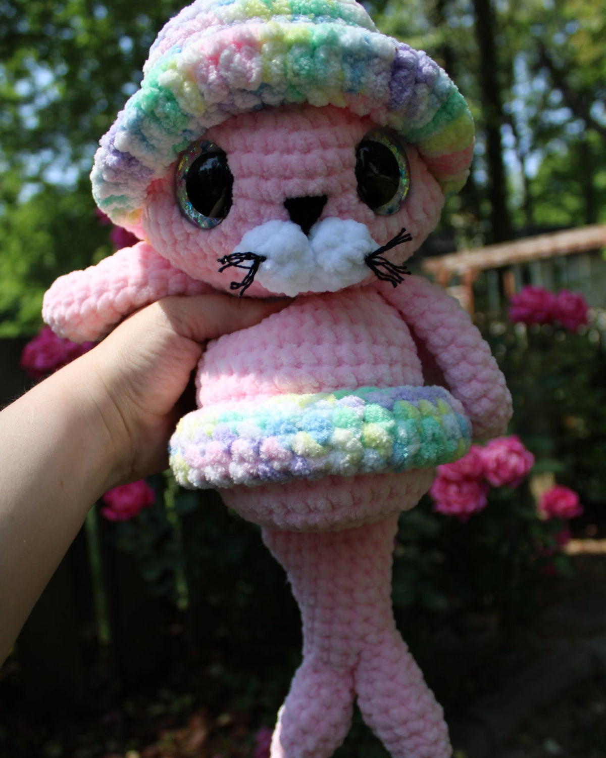 Crocheted pink seal plush toy with colorful hat held against a blurred natural background
