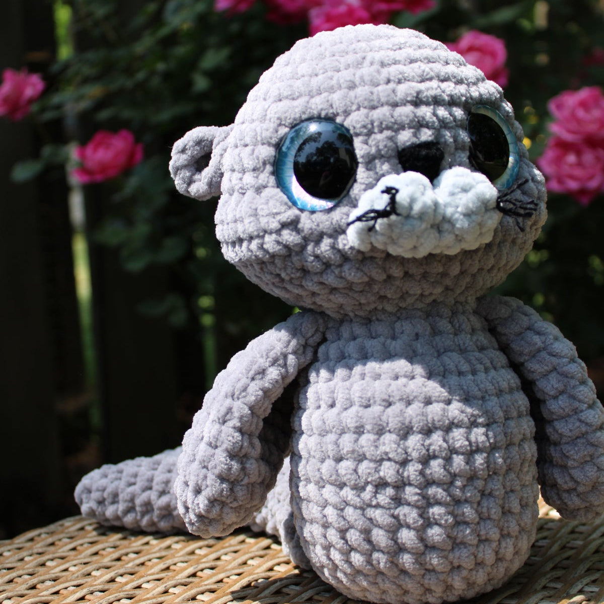 Crocheted seal toy on a wicker surface with pink flowers in the background