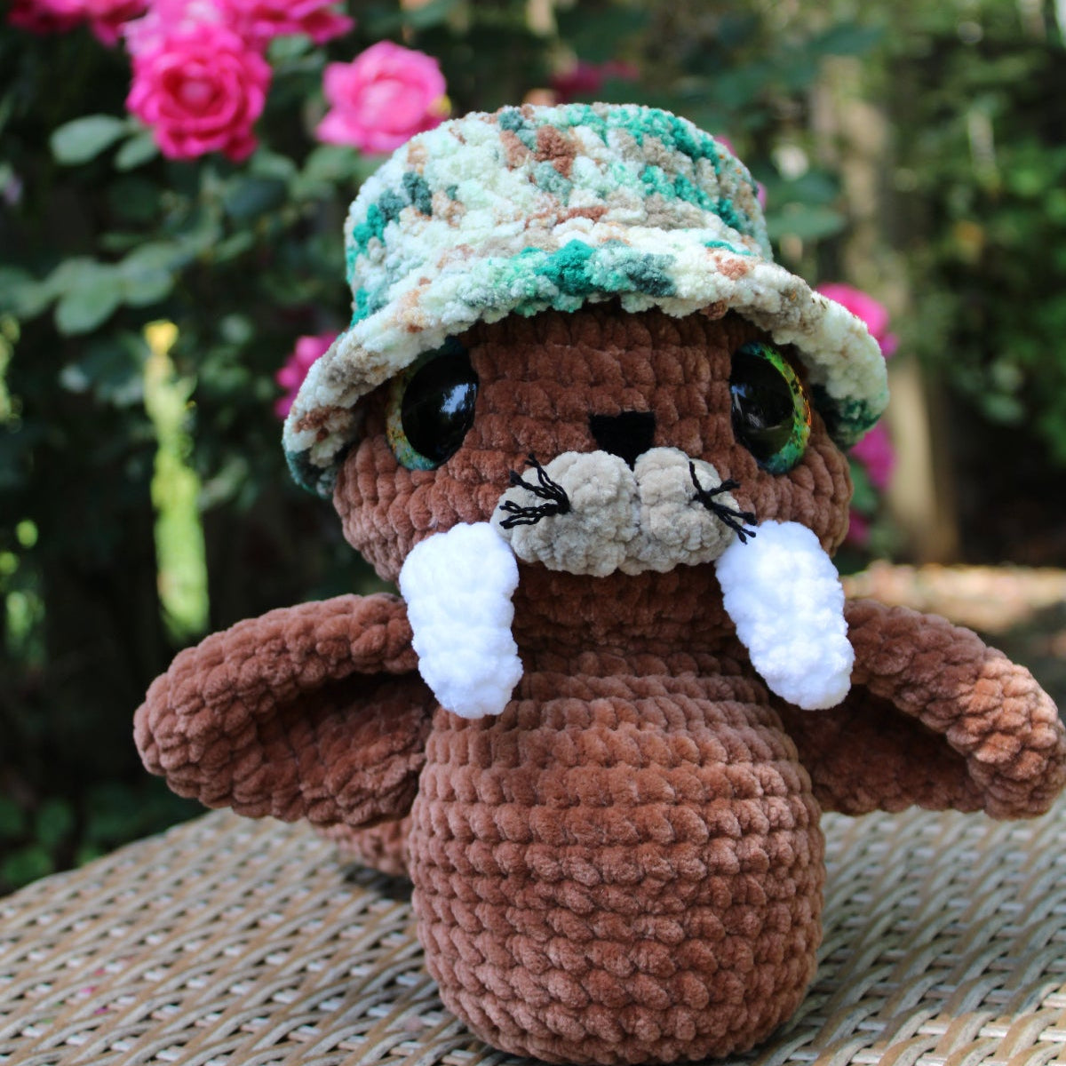 Crocheted animal figure with a hat and white accents sitting on a woven surface with pink flowers in the background.