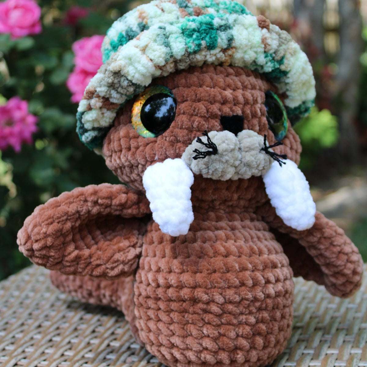 Crocheted animal plush toy with a colorful hat on a woven surface with flowers in the background