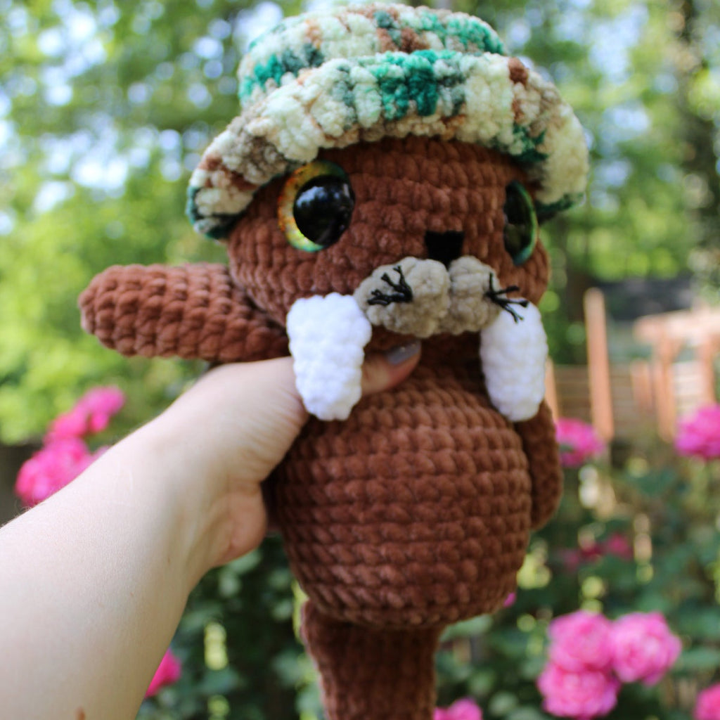 Hand holding a crochet walrus toy with a hat in front of a blurred natural background